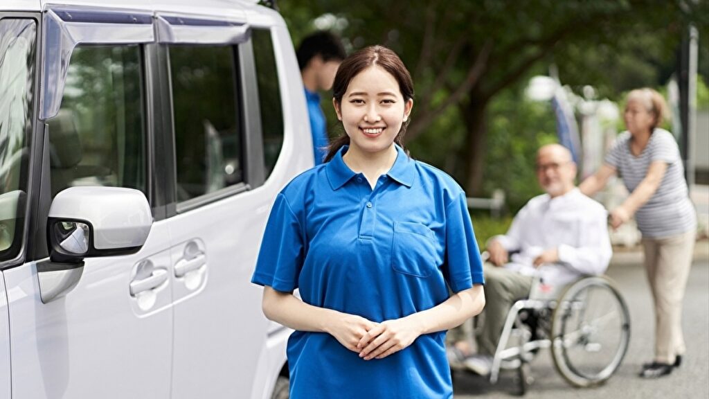 青いポロシャツを着て車の横に立つ介護士の女性